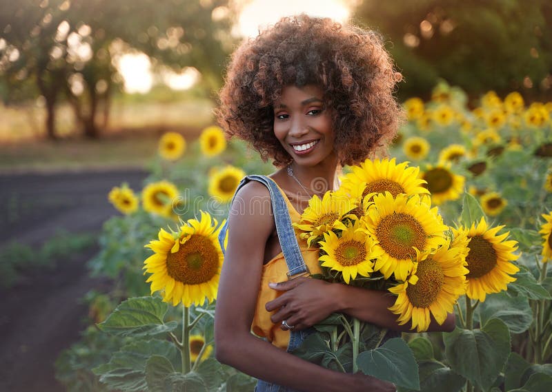 Happy young black woman walks in the sunflower field. Smiling dark-skinned girl with with a bouquet of sunflowers and