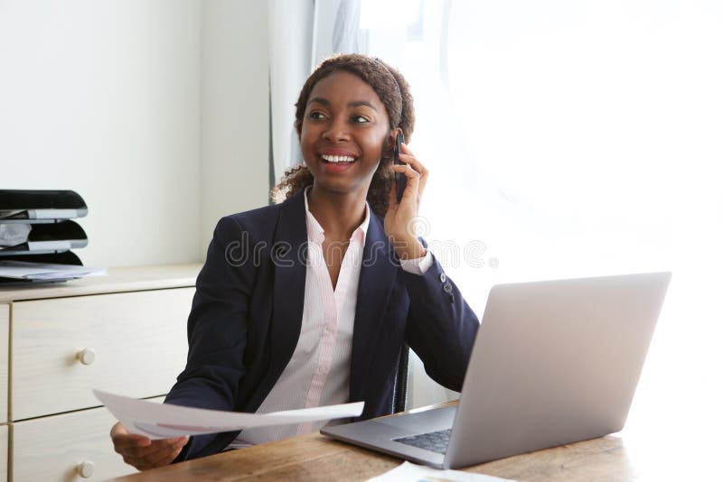 Happy young business woman sitting at office desk talking on mobile phone with a document in hand