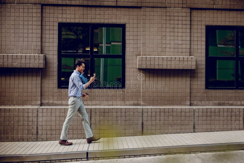 Happy Young Businessman in Casual wear Using or reading Mobile Phone while Walking by the Urban Building. Lifestyle of Modern People. Side View. Full Length