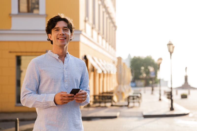 Happy young casual man holding mobile phone while walking at the city street