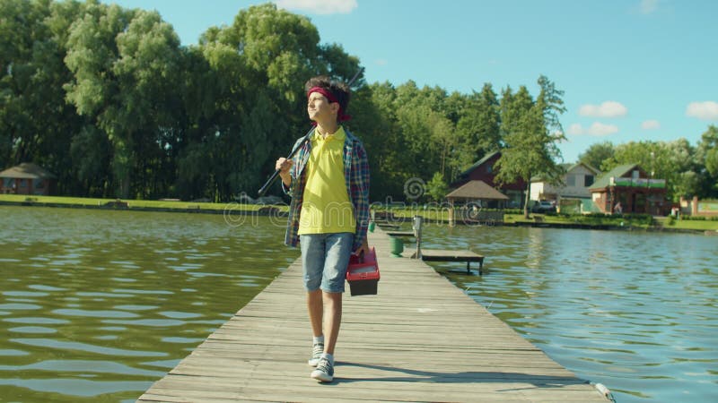 Happy young fisherman with fishing rod and tackle box going for angling on pond