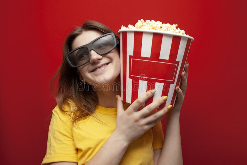 Happy young girl the viewer in 3D glasses holds a big box of popcorn and watches a movie on a red color background