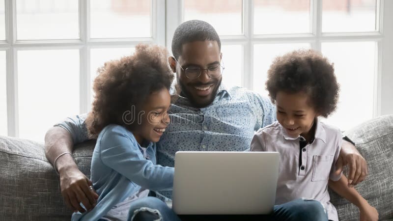 Happy young handsome african american father in eyeglasses embracing small adorable biracial kids, enjoying playing online game on computer or watching funny comedian movie together at home. Happy young handsome african american father in eyeglasses embracing small adorable biracial kids, enjoying playing online game on computer or watching funny comedian movie together at home.