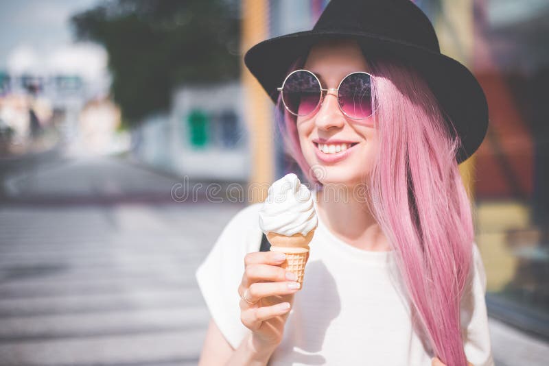 Happy young hipster woman with long pink hair, hat and sunglasses eating ice cream outdoors royalty free stock photography Happy young hipster woman with long pink hair, hat and sunglasses eating ice cream outdoors royalty free stock photography
