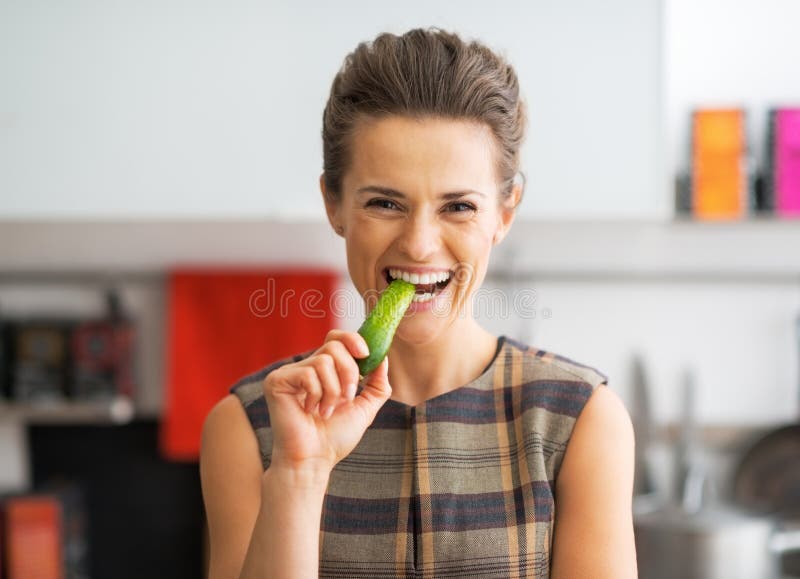 Happy Young Housewife Eating Cucumber In Kitchen Stock Photo