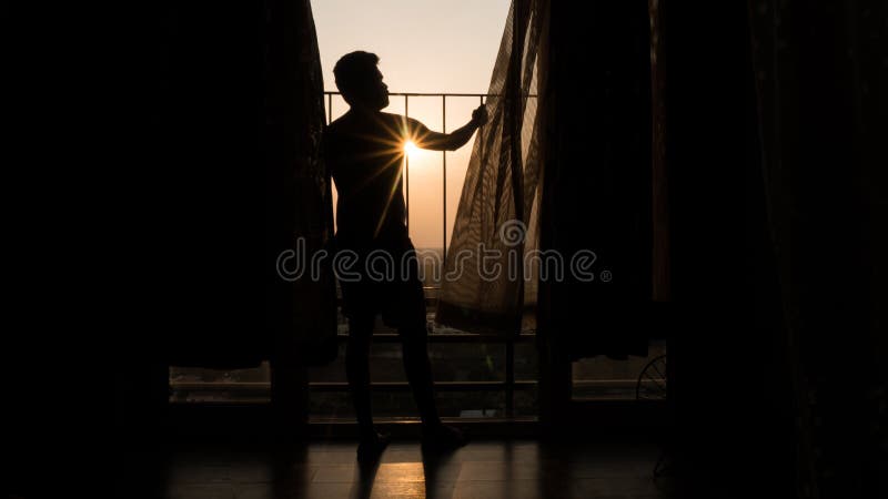 Happy young man enjoying a beautiful sunset from his apartment balcony with sun rays passing through his shoulders.