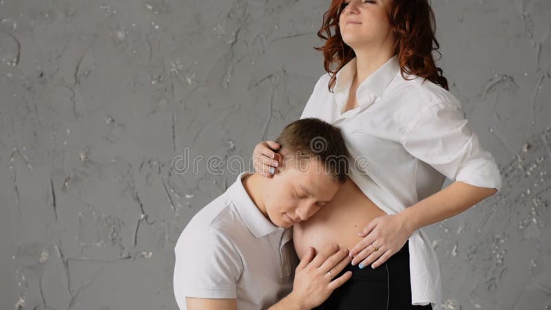 Happy young man nuzzles the belly of his pregnant wife. Young wife pats him on the head and look at the camera. stock video Happy young man nuzzles the belly of his pregnant wife. Young wife pats him on the head and look at the camera. stock video
