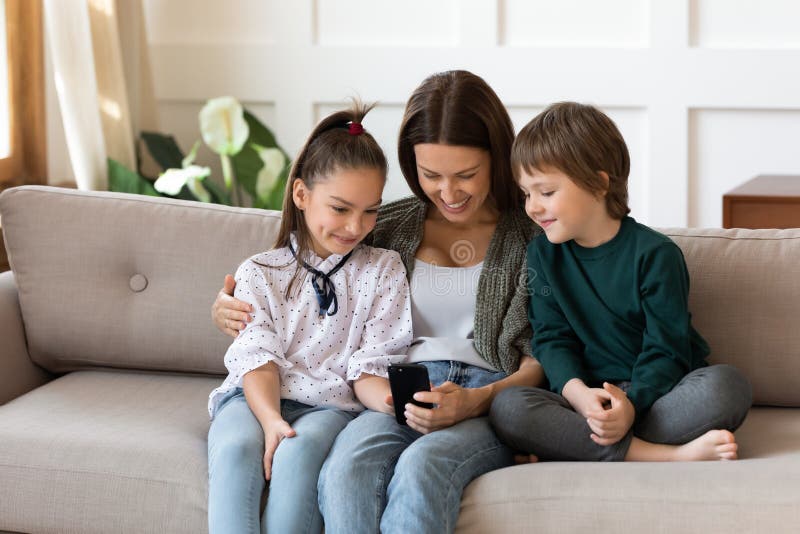 Happy Young Mother Embracing Daughter Son Looking At Smartphone Screen