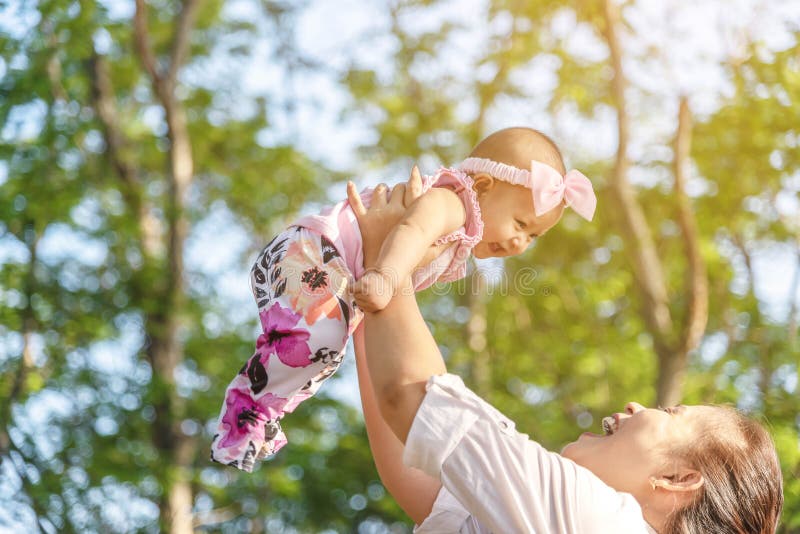 Happy young mother playing with little 5 months daughter in park. Lovely baby girl laughing while mother holding her in the air