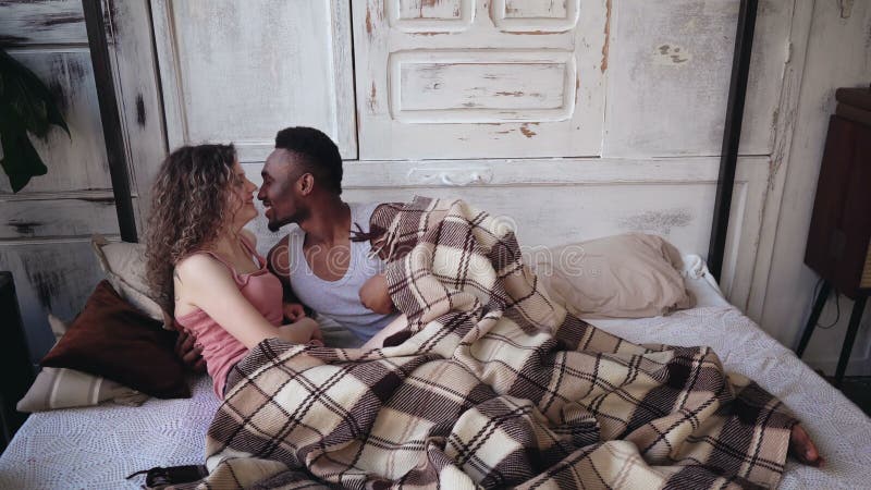 Happy young multiethnic couple lying under blanket, hugging and kissing. Tenderly morning of man and woman on the bed. stock photo Happy young multiethnic couple lying under blanket, hugging and kissing. Tenderly morning of man and woman on the bed. stock photo