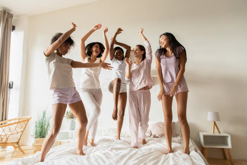 Happy young multiracial women in pajamas jumping on bed.
