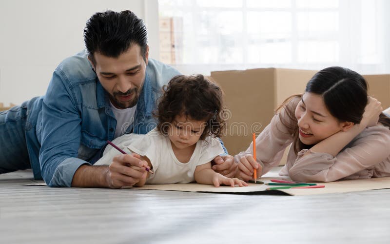 Happy young parents Caucasian father and Asian mother lying on floor and play drawing with little kid girl at home.