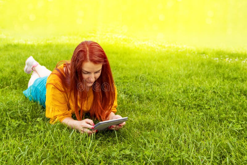 Young red-haired girl in a yellow jacket and green skirt lying on the grass and using a tablet, watching an educational video in a stock images Young red-haired girl in a yellow jacket and green skirt lying on the grass and using a tablet, watching an educational video in a stock images