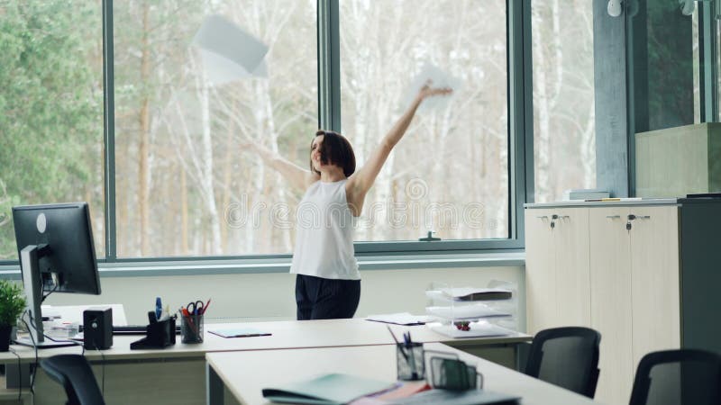 Happy young woman is dancing in office taking off jacket and throwing papers tossing her hair enjoying freedom and good