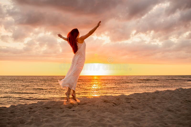 Happy young woman in a long white dress looking at the sunset on empty sand beach with her hands up. Freedoom, vacation