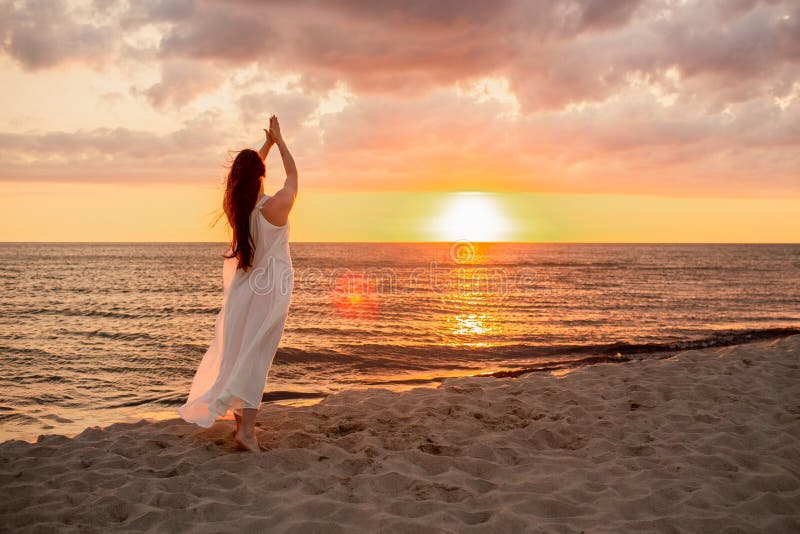 Happy young woman in a long white dress looking at the sunset on empty sand beach with her hands up. Freedoom, vacation