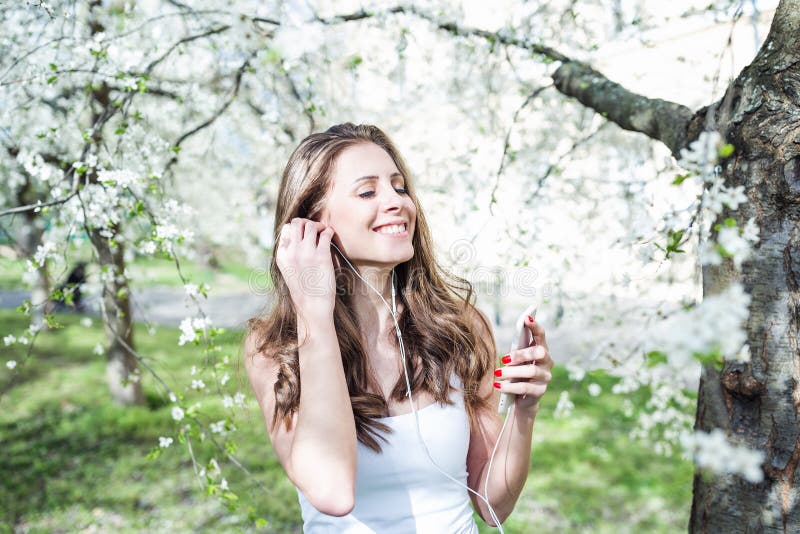 Young woman smiling and with closed eyes listening to music on the phone with earphones under the blooming trees royalty free stock photo Young woman smiling and with closed eyes listening to music on the phone with earphones under the blooming trees royalty free stock photo