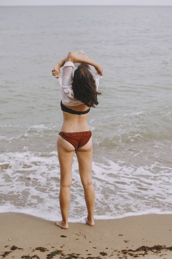 Happy young woman taking off wet white shirt on beach, back view. 