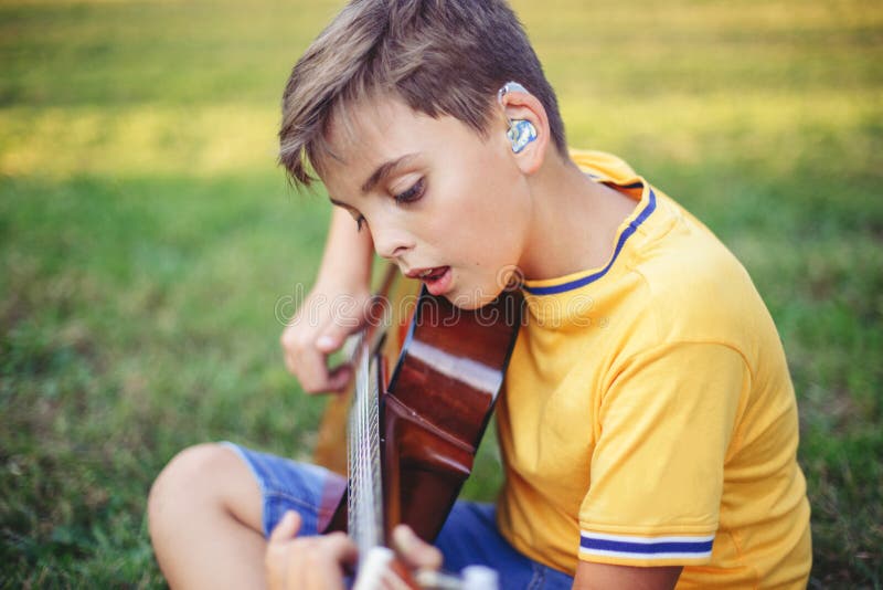 Hard of hearing preteen boy playing guitar and singing. Child with hearing aids in ears playing music and singing song in a park. Hard of hearing preteen boy