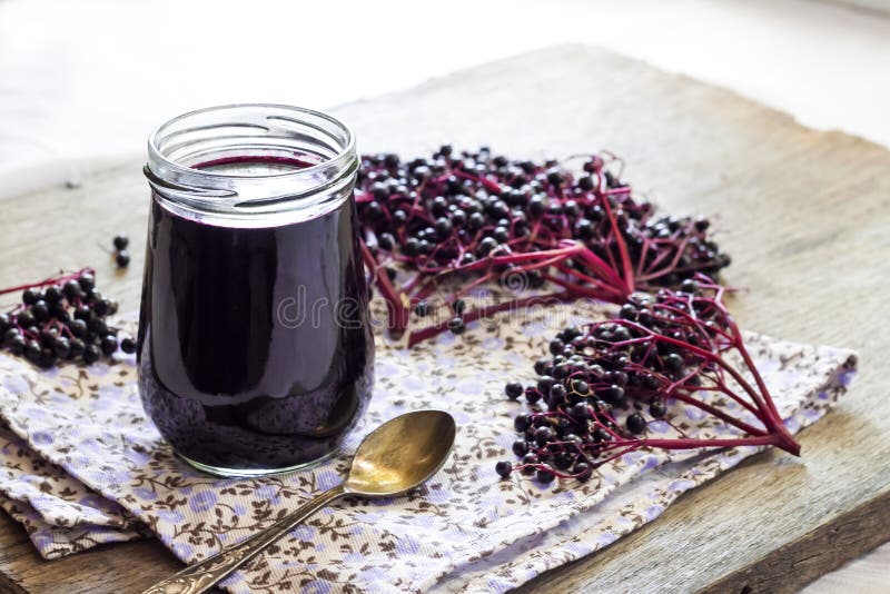 Homemade black elderberry syrup in glass jar and bunches of black elderberry in background. Homemade black elderberry syrup in glass jar and bunches of black elderberry in background