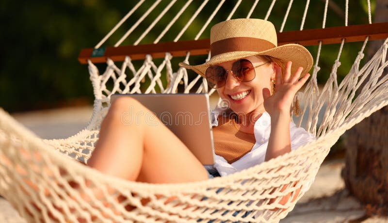 Happy young female wearing straw hat waving while having video call on tablet pc relaxing in the hammock on tropical beach