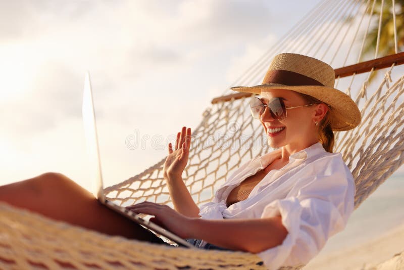 Happy young female wearing straw hat waving while having video call on laptop, relaxing in the hammock on tropical beach