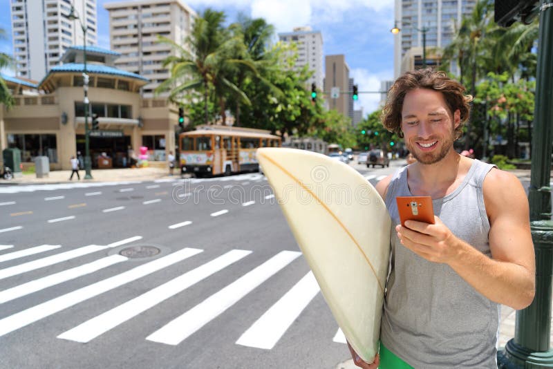 Hawaii surfer man Honolulu city lifestyle using smart phone going to Waikiki beach surfing holding surfboard on street. American living in USA. Hawaii surfer man Honolulu city lifestyle using smart phone going to Waikiki beach surfing holding surfboard on street. American living in USA