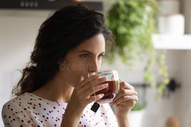 Head shot close up of beautiful woman drinking black tea, enjoying morning, breakfast, starting new day, young female holding glass cup with tea bag inside, hot beverage, relaxing during break. Head shot close up of beautiful woman drinking black tea, enjoying morning, breakfast, starting new day, young female holding glass cup with tea bag inside, hot beverage, relaxing during break