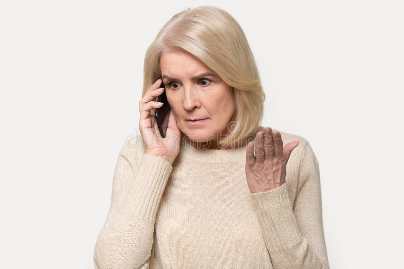 Headshot of annoyed old woman having argument talking via smartphone. Angry aged female speak irritated by cell phone, find out bad news over telephone. Portrait of lady with mobile isolated on grey