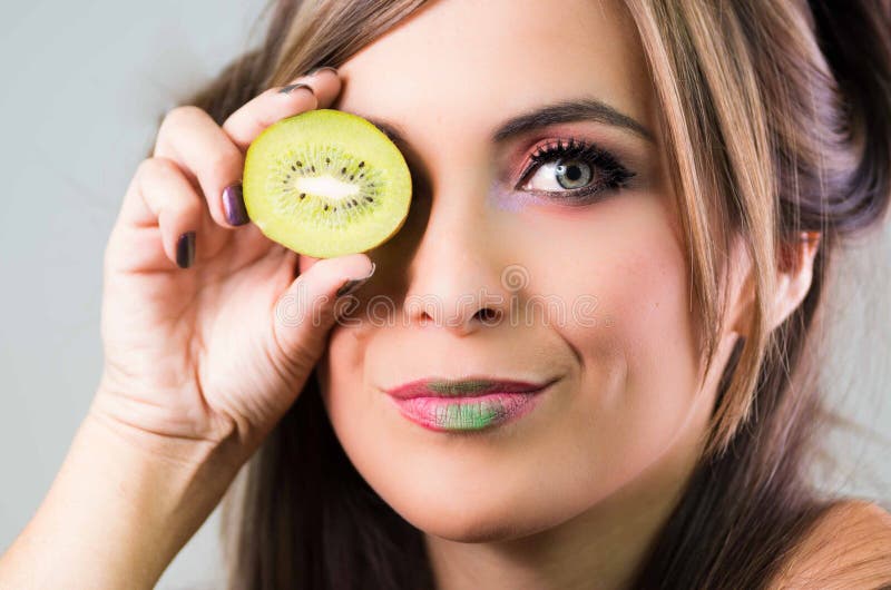 Headshot brunette, dark mystique look and green lipstick, covering one eye with open kiwi, looking into camera stock photos