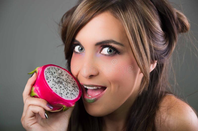 Headshot brunette, dark mystique look and green lipstick, holding open pink pitaya fruit next to mouth while sticking stock images