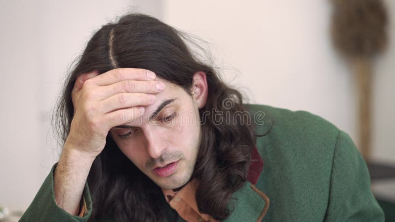 Headshot of frustration young snygg brunette man med långt hår och bruna ögon som dricker kaffe och som sitter i lager videofilmer
