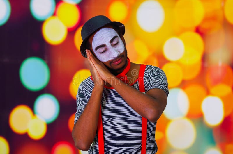 Headshot pantomime man with facial paint posing for camera using hands interacting sleeping, blurry lights background royalty free stock images