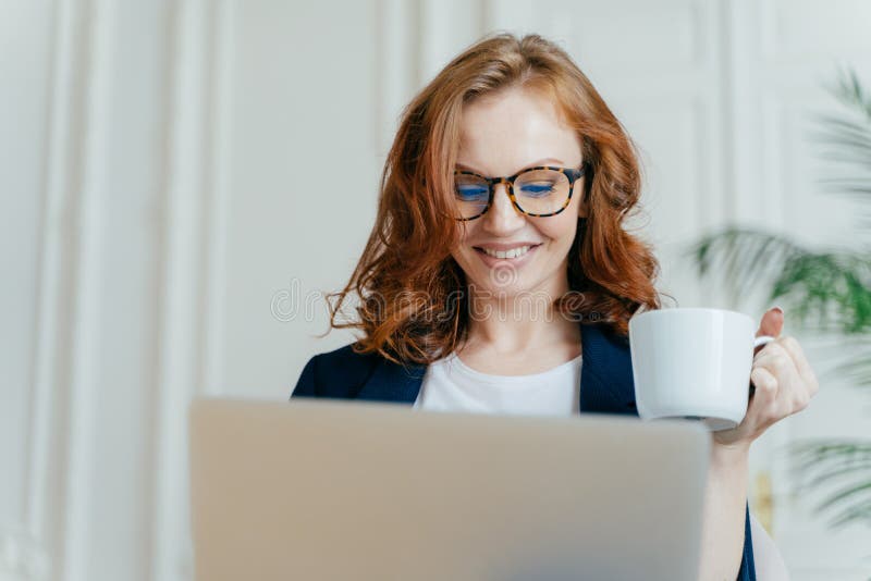 Headshot of smiling red haired female in optical glasses, enjoys hot coffee or tea, focused in laptop computer, makes purchases royalty free stock images Headshot of smiling red haired female in optical glasses, enjoys hot coffee or tea, focused in laptop computer, makes purchases royalty free stock images