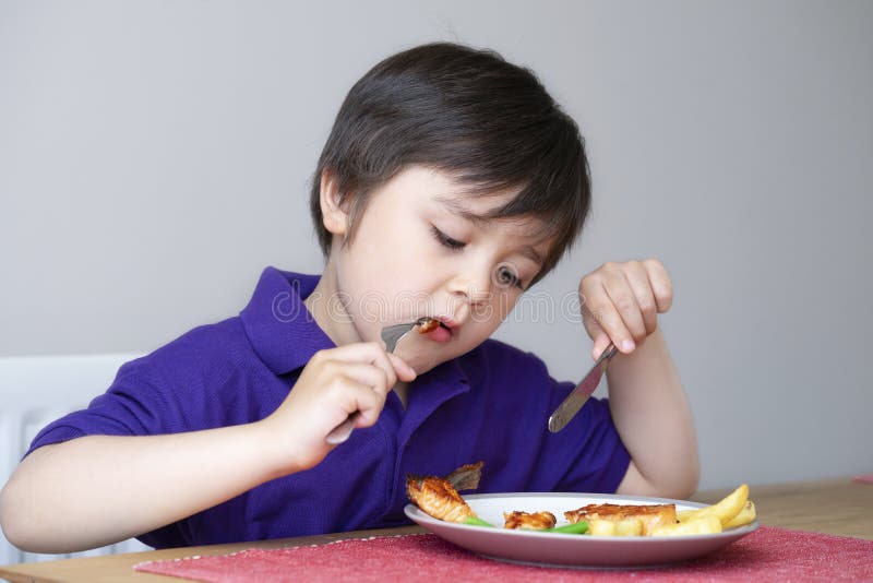 Healhty kid eating homemade salmon steak and chips for his sunnday dinner at home, Portrait of Child boy learning how to use knife