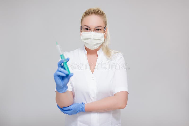 Health care and vaccination concept - female doctor or nurse in white uniform and mask holding syringe over gray