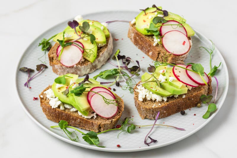 Healthy avocado toasts with radish, feta cheese, pea sprouts and pepper in a plate on white marble table for healthy diet breakfast