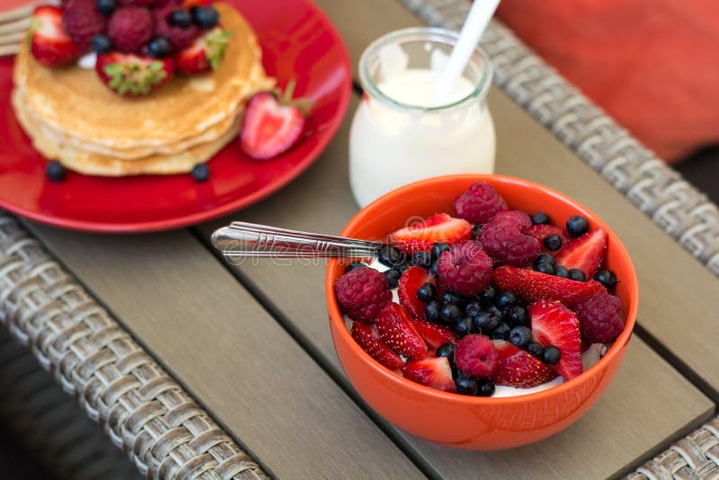 Healthy breakfast on garden furniture: cottage cheese with sour cream, strawberry, raspberry and blueberry, pancakes, yogurt