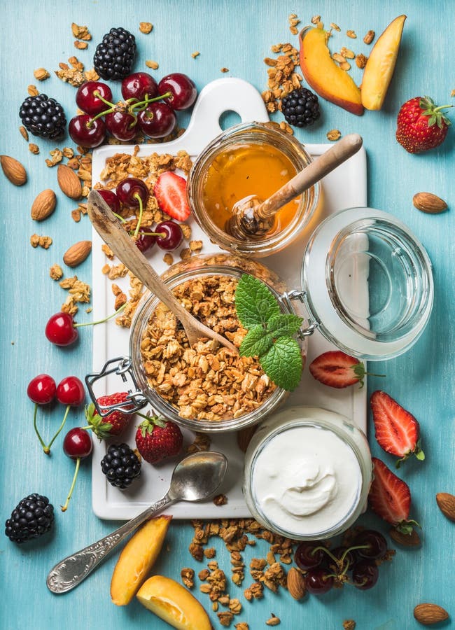 Healthy breakfast ingredients. Oat granola in open glass jar, yogurt, fruit, berries, honey and mint on blue background royalty free stock image Healthy breakfast ingredients. Oat granola in open glass jar, yogurt, fruit, berries, honey and mint on blue background royalty free stock image