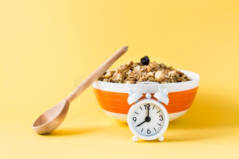 Healthy eating. Alarm clock in front of a baked granola made from oats, nuts and raisins in a bowl and a wooden spoon stock image