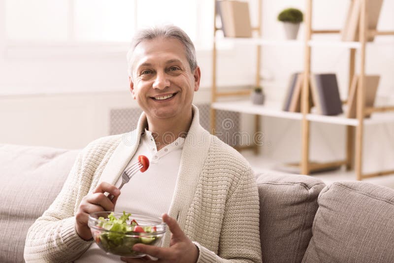 Healthy eating. Happy senior man eating fresh vegetable salad, sitting on sofa at home