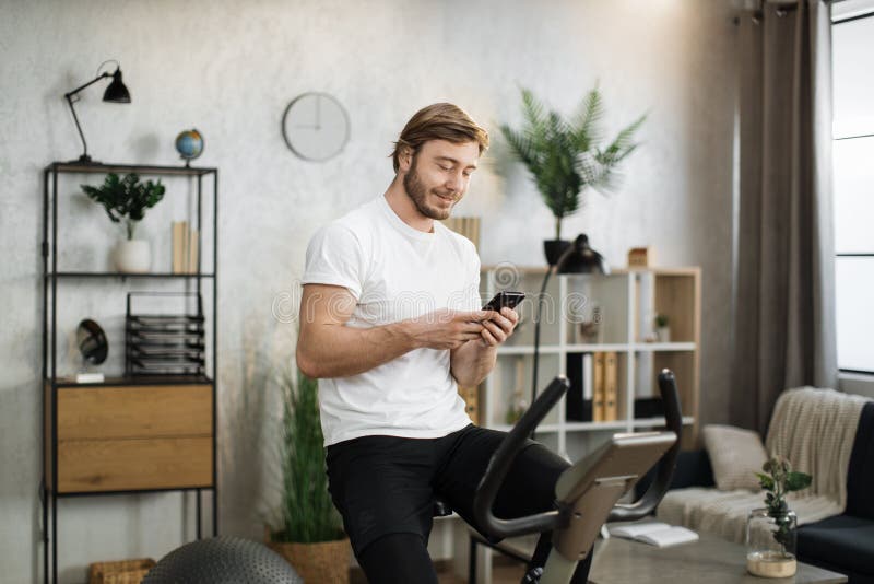 Healthy fit smiling asian man training at home on exercise static bike during workout holding phone