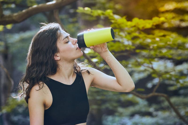 Healthy fitness girl drinking water from green bottle in forest