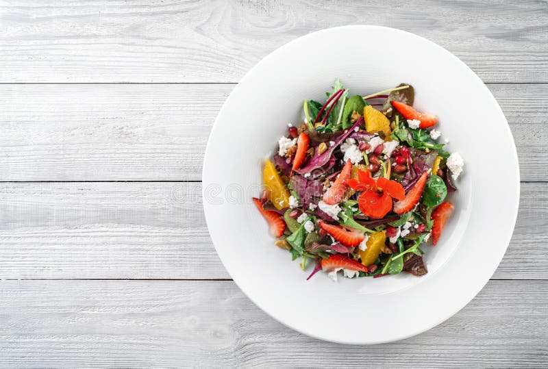 Healthy fresh salad with mix greens, strawberry, orange, nuts, cream cheese and edible flowers in plate on light wooden background stock image Healthy fresh salad with mix greens, strawberry, orange, nuts, cream cheese and edible flowers in plate on light wooden background stock image