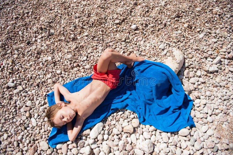 Healthy little kid in red shorts lying on blue towel on beach in hot summer day