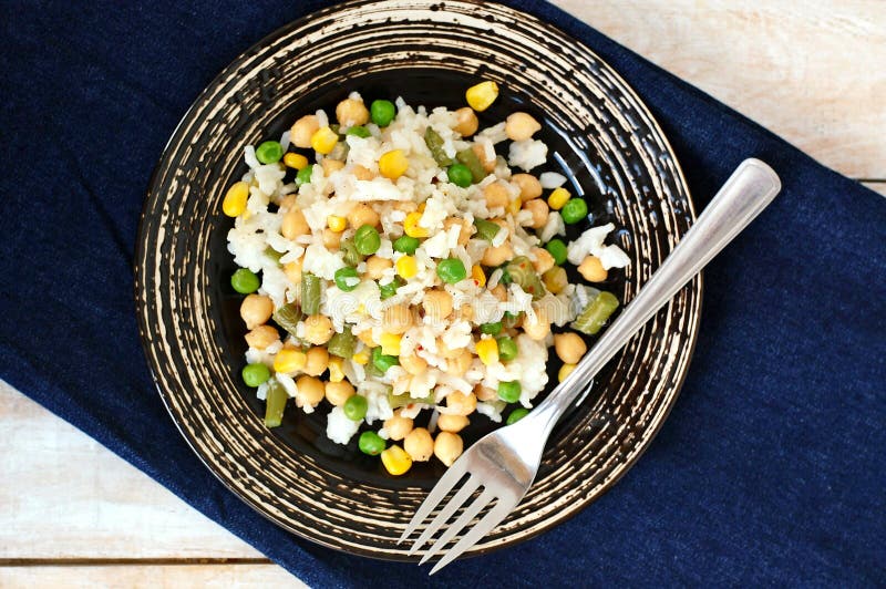 Healthy vegan meal with rice, chickpeas, sweet corn, peas, green beans and rice on dark plate on jeans cloth on wooden background