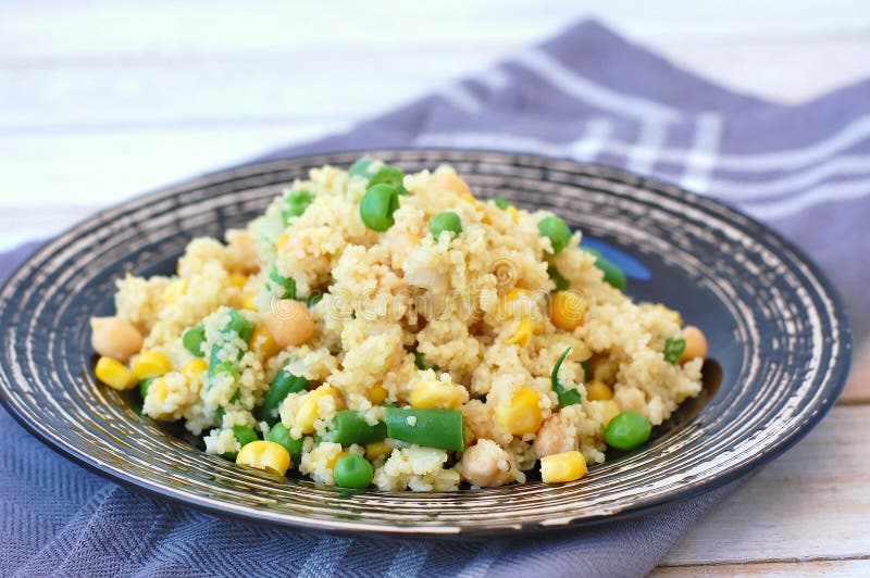 Healthy vegan meal with whole grain couscous, chickpeas, sweet corn, peas, green beans on dark plate on grey cloth on wooden