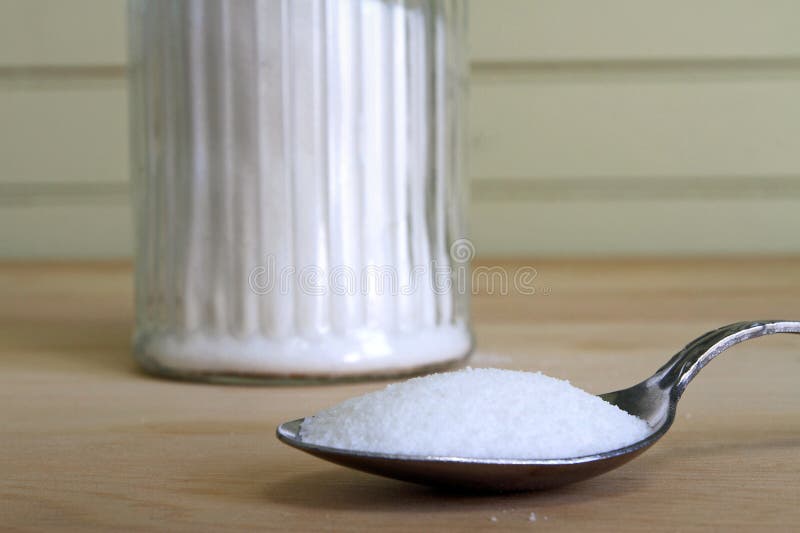 Heaping spoonful of sugar with glass sugar jar in background.