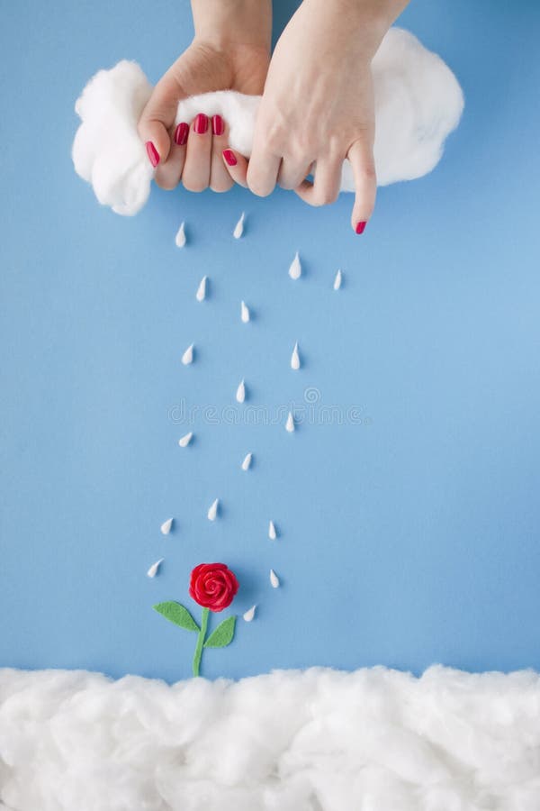 Two hands squeezing rain from a cloud over a tiny handmade red flower, in a heavenly scenery with clouds made of cotton-wool, on a blue sky paper background. Two hands squeezing rain from a cloud over a tiny handmade red flower, in a heavenly scenery with clouds made of cotton-wool, on a blue sky paper background.