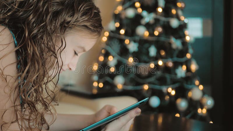 Heerlijk klein meisje speelt op een tablet op de achtergrond de kerstboom belicht met kerst.